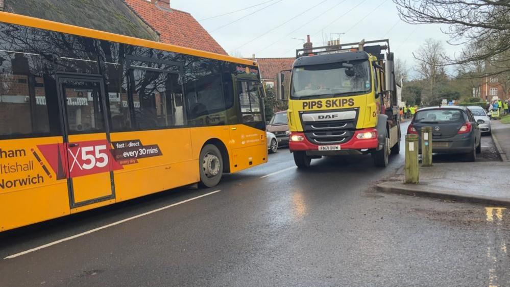 A yellow bus, branded the X55, and a yellow and red skip lorry face each other on the B1150 in Coltishall. There are parked cars on either side of the road, and a pavement build-out into the road to make for a pedestrian crossing point.