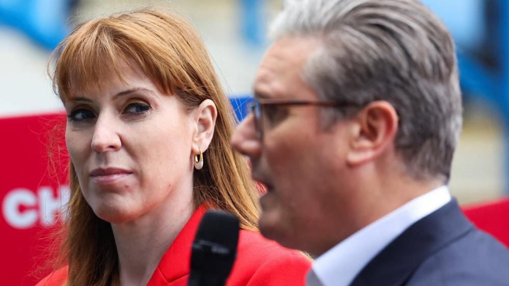 Angela Rayner stands next to Keir Starmer, and looks sideways at him as he speaks