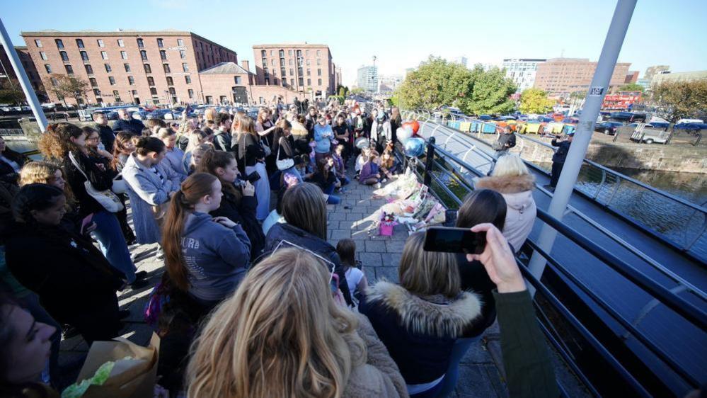 A crowd of people can be seen gathered on Keel Warf Bridge at Royal Albert Dock in Liverpool. They are looking at a collection of tributes, messages and flowers, left to Liam Payne on one side of the bridge.