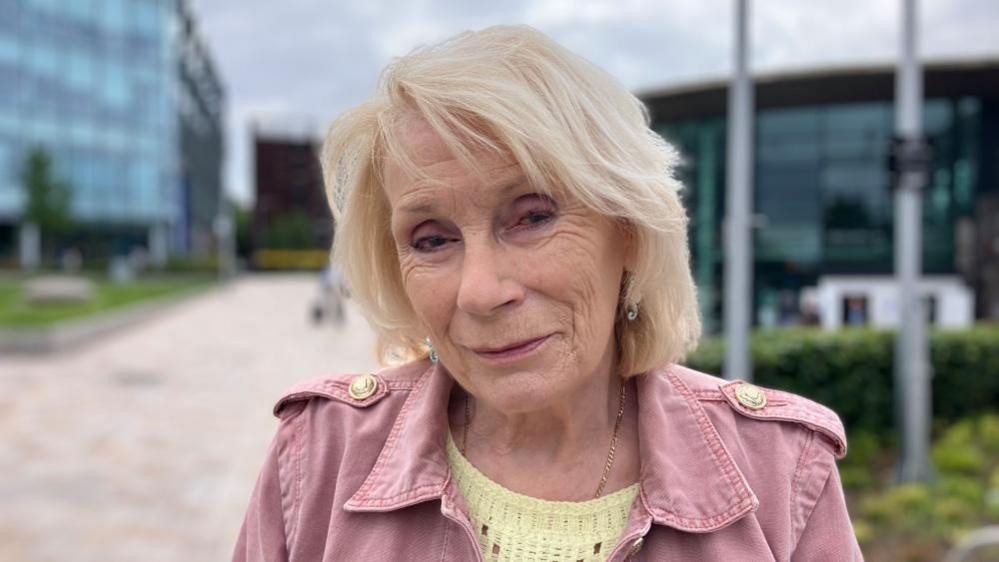 An older woman with a blond bob and wearing a yellow jumper and a pink jacket stands on a street.