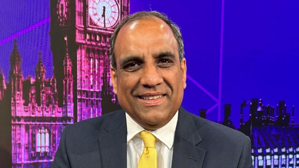 Shaffaq Mohammed, leader of the Liberal Democrats in Sheffield, sits in front of a digital backdrop showing Big Ben in London. He is smiling, wearing a dark suit, and yellow tie.