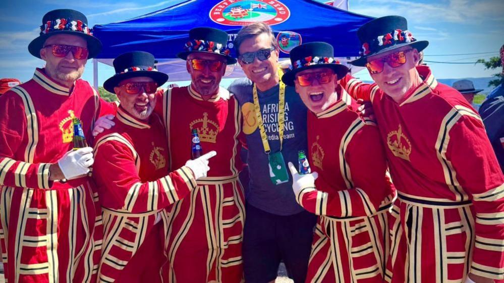 A group of men dressed up as British beefeaters stand in a line smiling, with a member of a cycling group. They wear black top hats with ribbons around the rim, red tunics with a crown across the chest, and sunglasses.