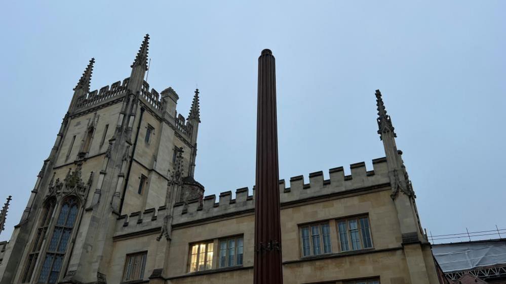 A lamp post, with a missing lamp, outside a building in Cambridge. The stone building is hold, has windows, ornate features, and the sky is blue. 