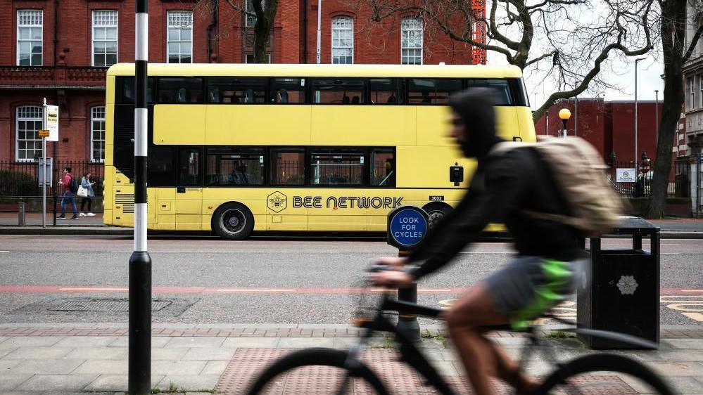 A man riding a bicycle is seen riding along a street. A yellow double-decker bus with the words "bee network" on the side is in the background. 