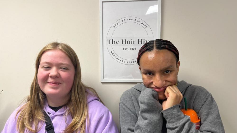 Molly with long blonde hair wearing a pink top and Cameron with short dark hair wearing a grey top. Cameron has his fists on his chin. They are sitting in front of a "Hair Hive" sign.