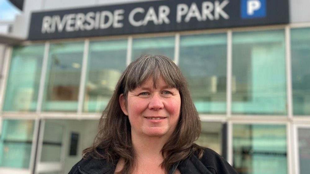 Sharon is wearing a black jacket behind and posing behind the Riverside Car Park.