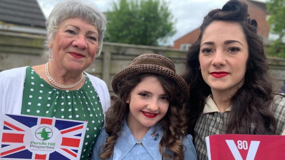 An older woman, a girl and a younger woman stand together smiling. They all have their hair and make-up done in a 1940s style and are also wearing retro dresses. They are holding VE Day and Union Jack flags.