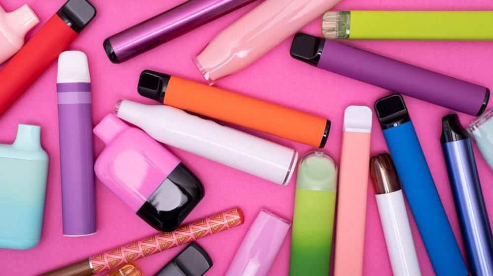 A stock image shows an assortment of disposable vapes strewn on a pink surface