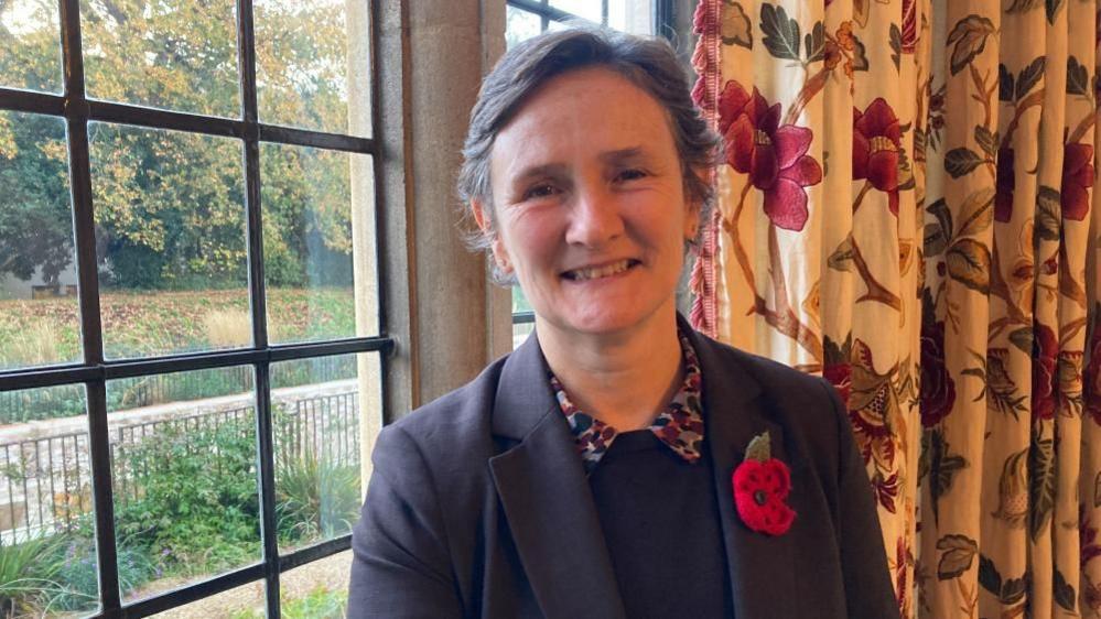A woman, wearing a smart jacket and a poppy is smiling at the camera.