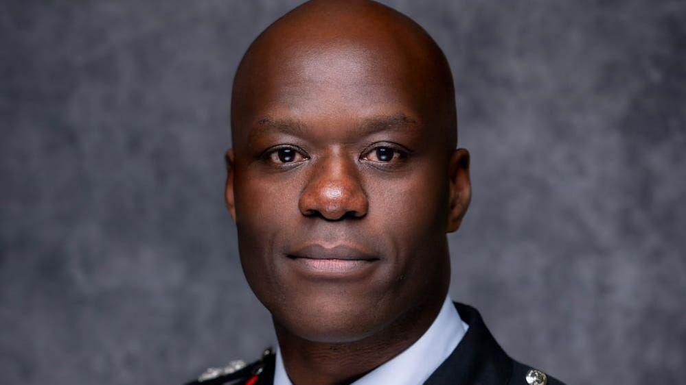 A man with a shaved head and smiling face is wearing formal uniform and looking at the camera in a publicity shot. He is standing against a dappled grey background.