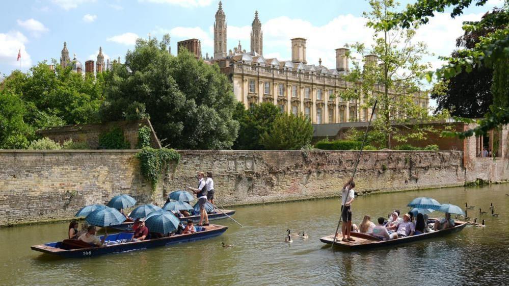 Punters in three punts are taken across the water next to a walled section of the river.