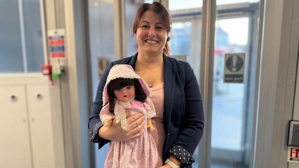 Pip Martin in a pink top and blue jacket, with her dark hair tied back, holding Annabelle the doll, which is in a pink dress and pink bonnet