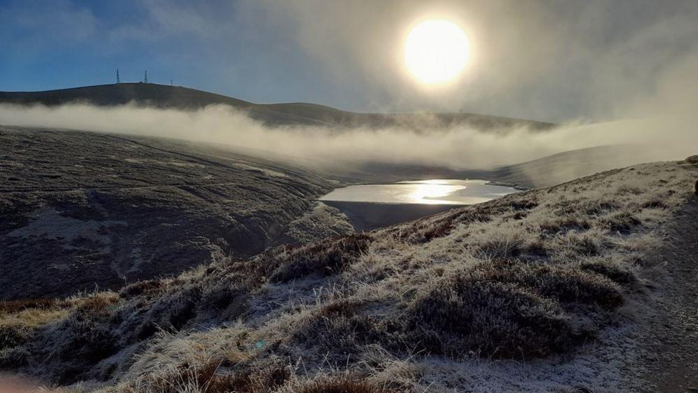 Frozen grassy hills with a light fog and a bright burning sun in a blue sky.