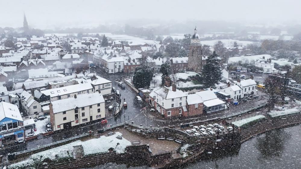 An aerial shot of Upton-upon-Severn with the roofs of buildings covered in snow. The river is in the foreground of the picture. 