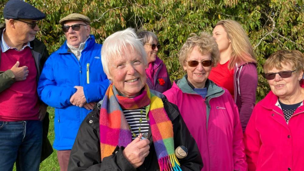 Jan Hupfield with short light-coloured hair wearing a multicoloured scarf and dark raincoat with other people around her standing on grass.