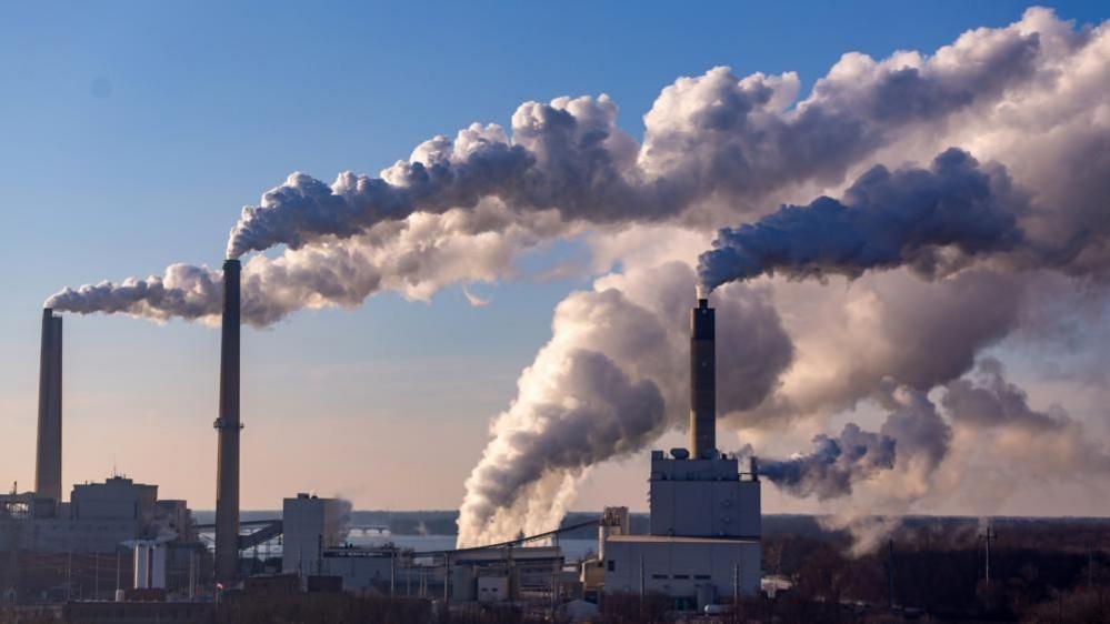 Wind is blowing clouds of pollution from the large smoke stacks of coal burning power plant. It is daytime and the sky is otherwise clear and bright blue.