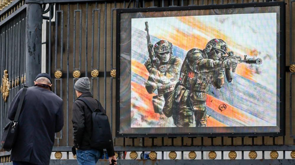 People walk past an electronic screen showing an image of Russian soldiers in downtown Moscow