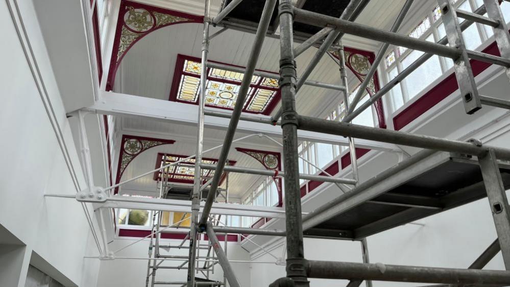 Cast iron brackets and timber, painted in white and deep plum colour, and stained glass windows form a lantern in the former department store. Scaffolding towers have been erected in the near ground and background of the image.