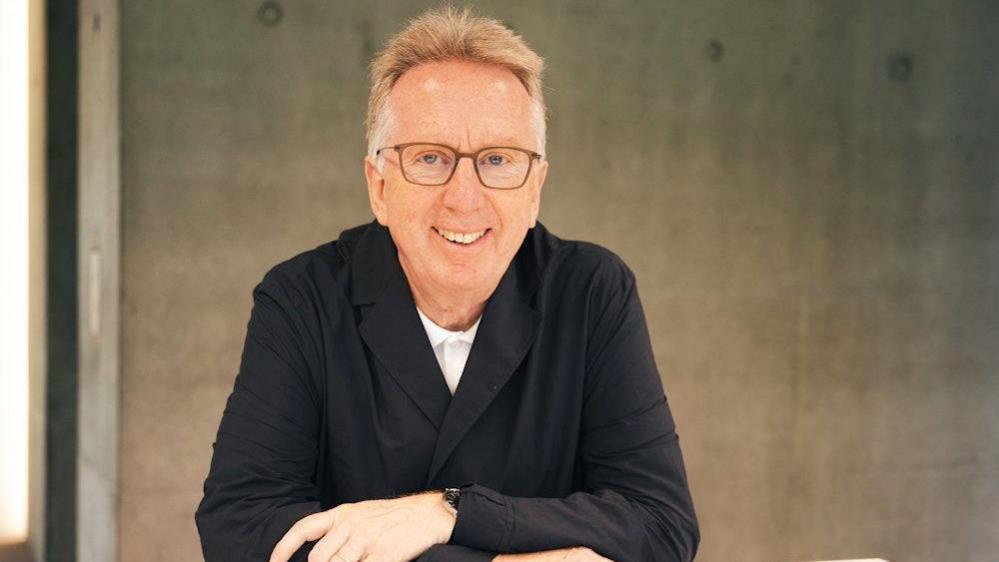 An image of Bob Allies sitting down with his arms folded and rested on a table in front of him. He is wearing a white collared shirt, a black blazer and brown framed glasses. He is smiling at the camera, and appears to be in an industrial type room with bare concrete walls.