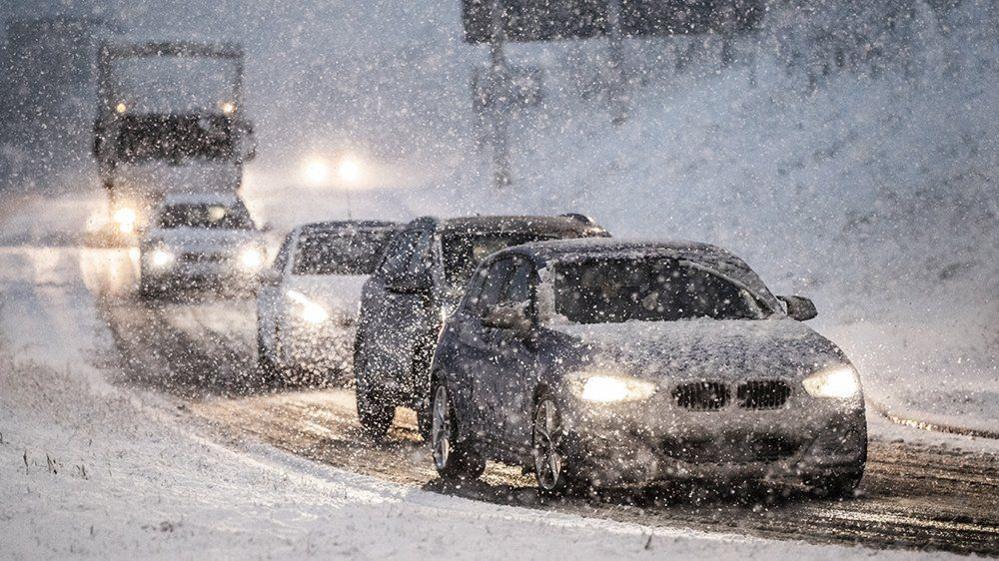 Cars and a truck drive slowly through heavy snowfall at Leeming Bar in North Yorkshire. The vehicles have their fog lights on, and are covered in snow. They sides of the road are also covered in snow.