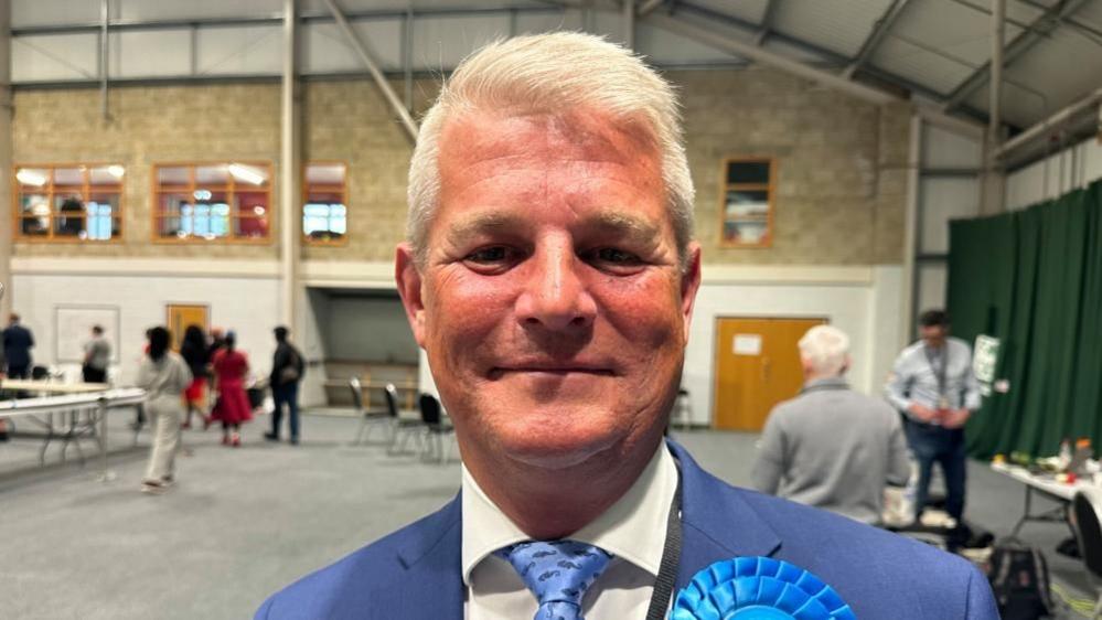 Stuart Andrew with short white hair wearing a blue suit and blue rosette