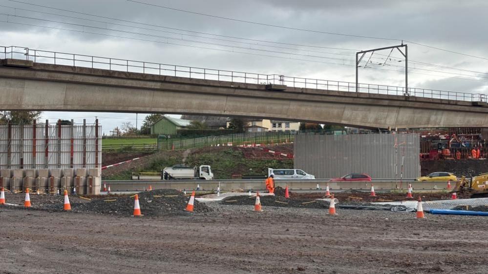 A large concrete bridge goes over a motorway. The ground underneath the bridge is all dug up and a line of traffic cones are placed along it. The area is cordoned off with tape.