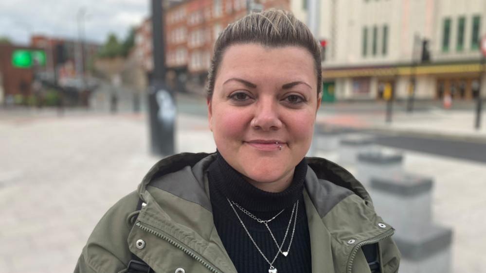 A woman wearing a camo jacket and black turtleneck stands smiling in Stockport's town centre.