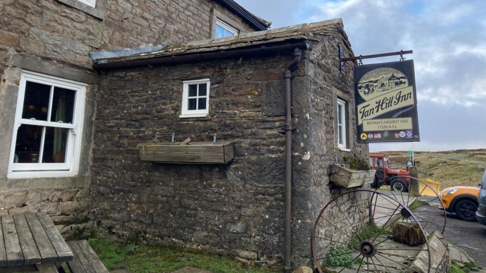 The pub is a beige stone building, with a blackboard style sign that welcoming customers to "Britain's Highest Inn at 1732 metres". In the background moorland, a tractor and a car can be seen.