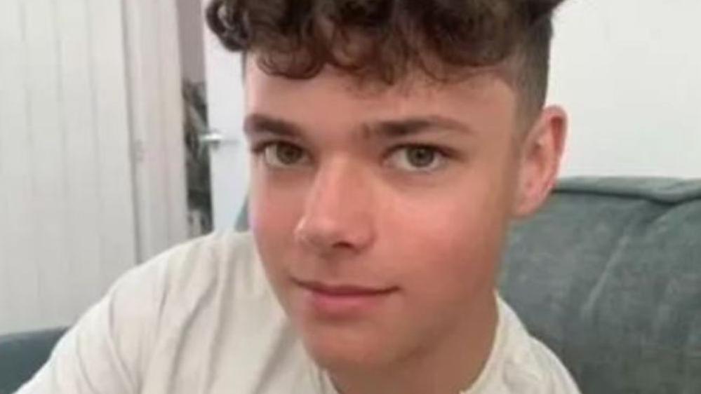 A teenage boy with brown curly hair, short on the sides. He is wearing a white Under Armour t-shirt and a thin chain as he sits on an armchair.