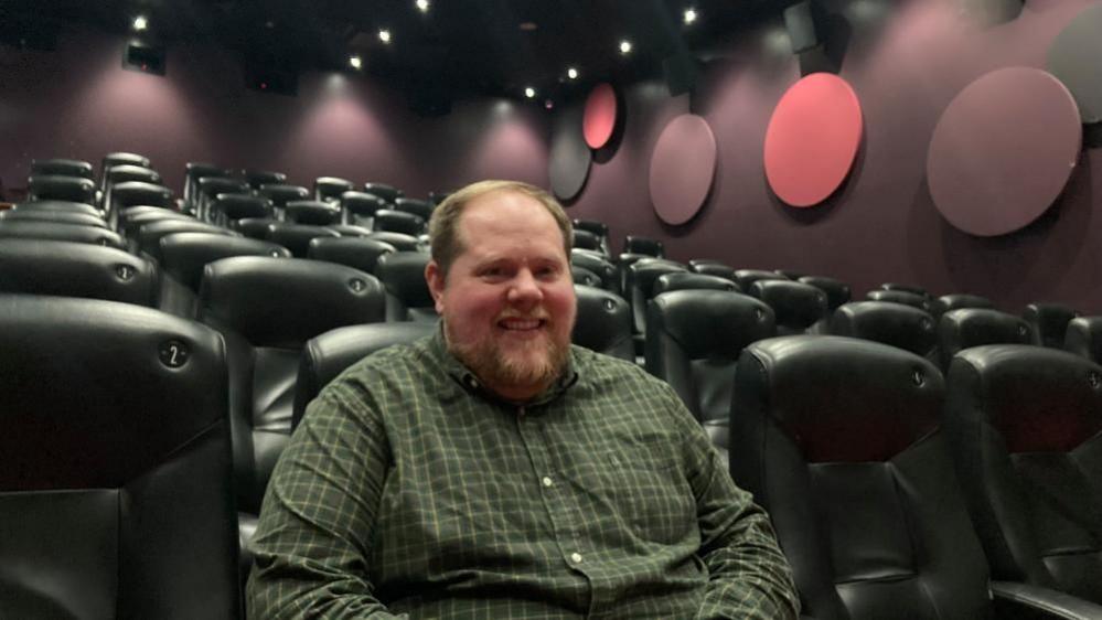 Richard Clifford sitting in a cinema, smiling and looking at the camera, and wearing a green shirt