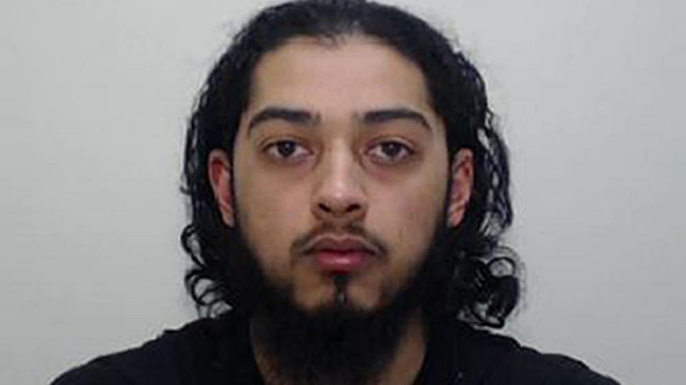 A mugshot of a young man with long dark hair and a dark beard. He has a blank expression on his face.