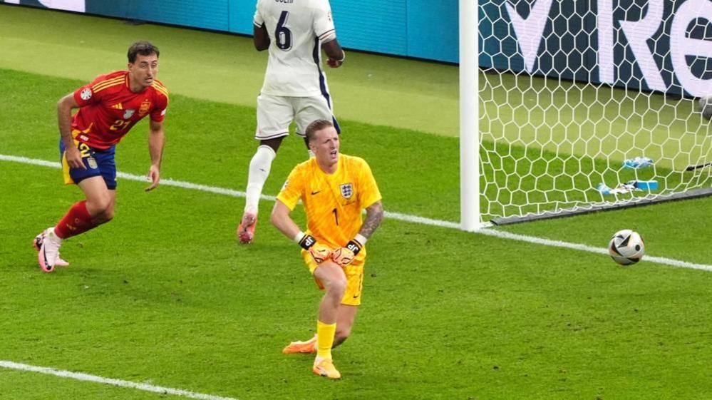 Mikel Oyarzabal with short dark hair has just kicked the ball past Jordan Pickford (in yellow) into the goal