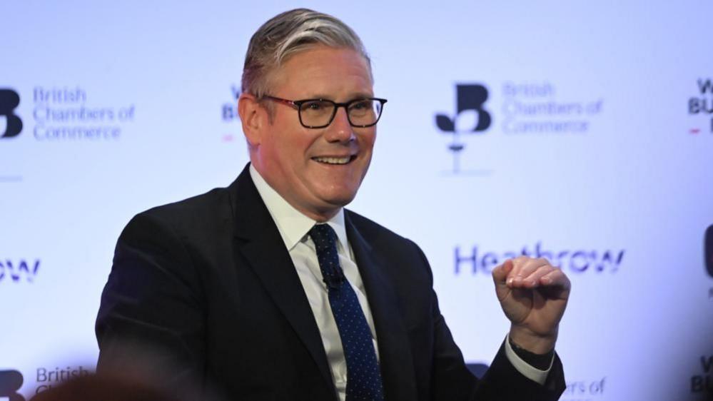 Sir Keir Starmer stands smiling in front of a white banner. He is holding one hand up in front of him.