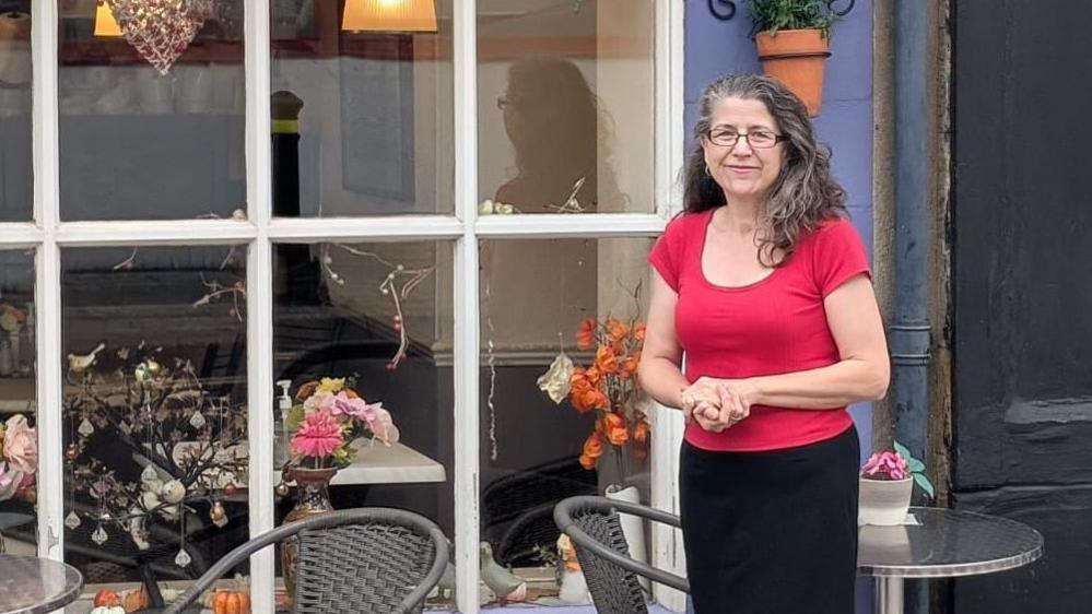 A woman standing outside her cafe in Yeovil. She has long dark hair and is wearing a red t-shirt and black bottoms. She is pictured outside, next to some tables and chairs.