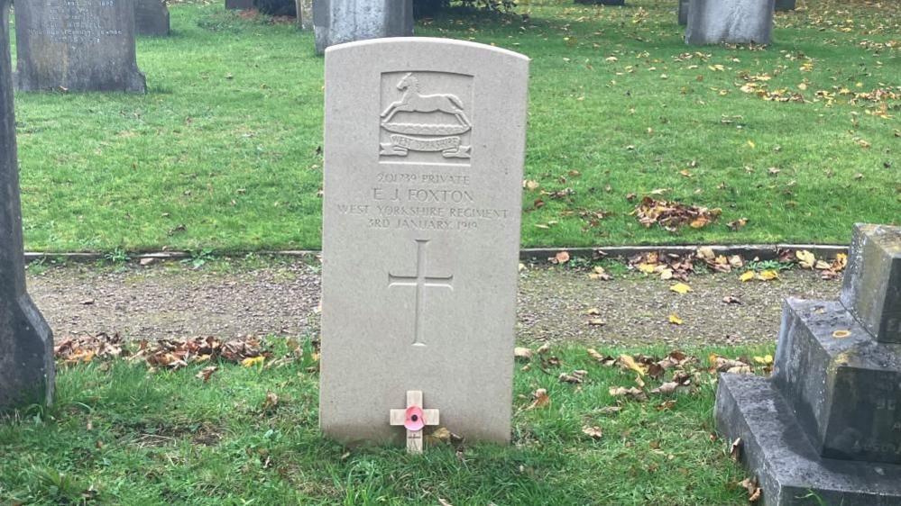 Emmanuel J. Foxton's Commonwealth War Grave in St Helen's churchyard. The grave is a light-coloured stone, bearing an engraving of a horse above a banner, Emmanuel J. Foxton's name, date of death and a cross. It is positioned next to an older stone cross headstone, in a row with other grey headstones and on a grassy area divided by a path.