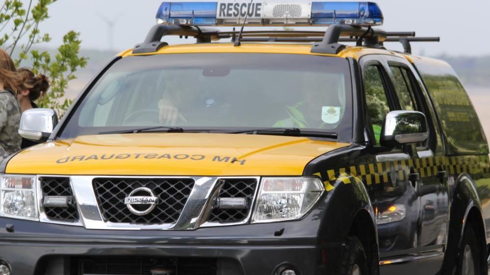 Two people sit in the front of a Nissan 4 by 4 vehicle with "HM Coastguard" written on the bonnet and blue lights on the roof, with the word "rescue".