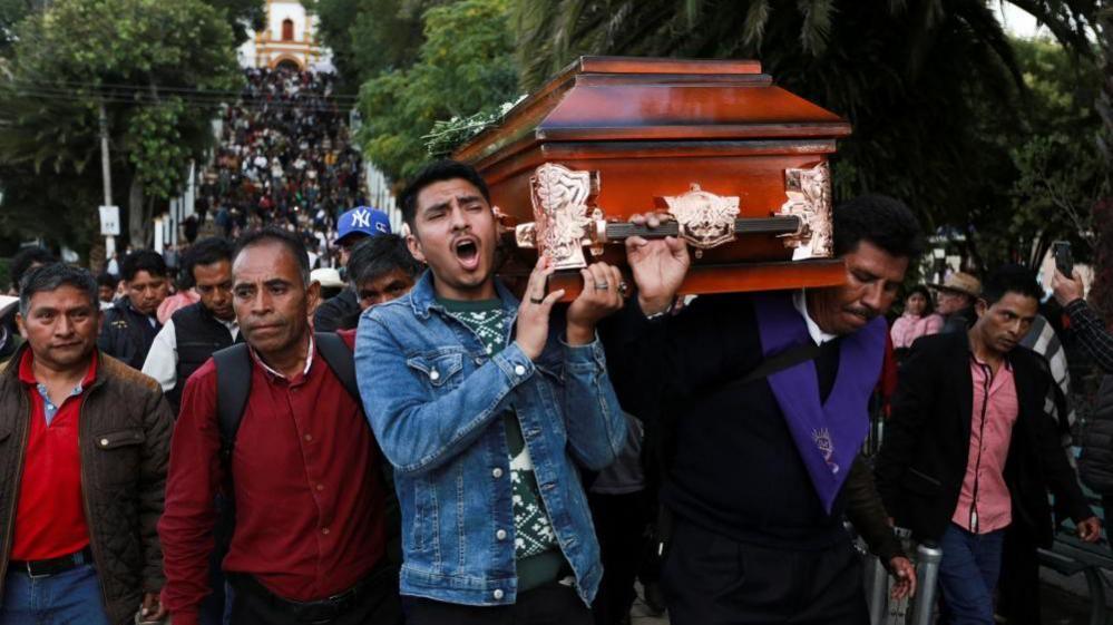 Men carry the coffin of Father Marcelo on their shoulders while surrounded by people mourning the priest. 