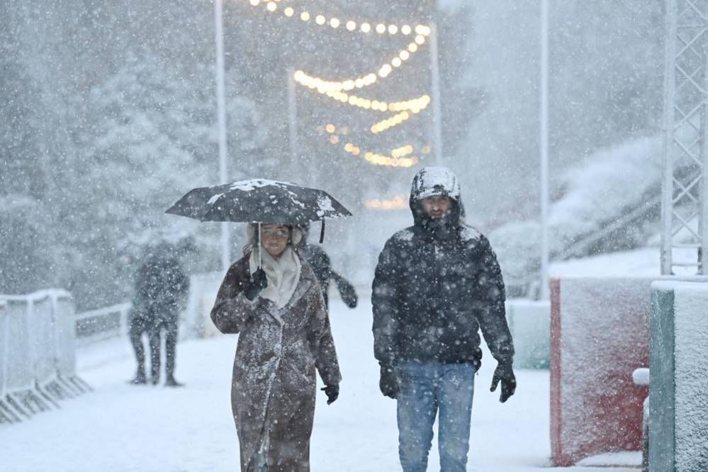 A woman, wearing a brown furry coat and cream scarf, while carrying an umbrella and a man wearing a black puffer jacket and jeans are covered in snow. Snow lying on the ground and falling from the sky is visible in the backround