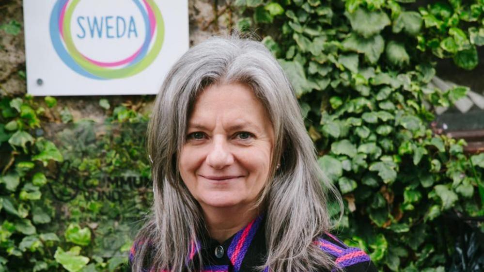 Paula wearing a pink and black top, with long grey hair standing in front of an ivy covered wall and a white sign with her charity's logo on it.