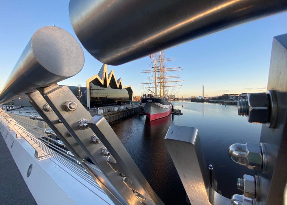 A view of Glasgow’s Riverside Museum and tall ship Glenlee on a sunny day, taken through the new Govan-Partick bridge. The River Clyde is still and there's a blue sky.
