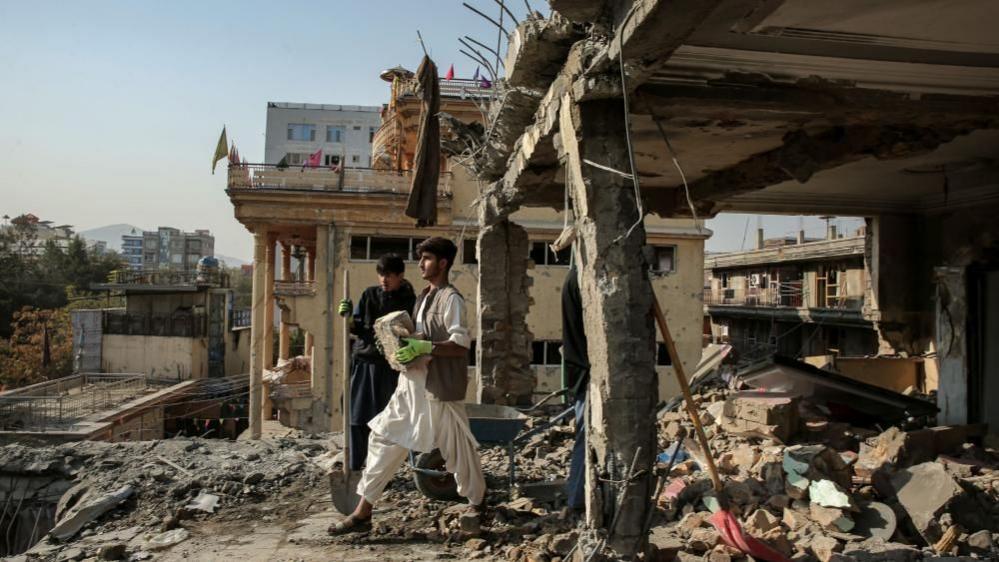 Two young men, one in all black holding a spade and another in white carrying a brick, stand amid the rubble of a destroyed building in Kabul