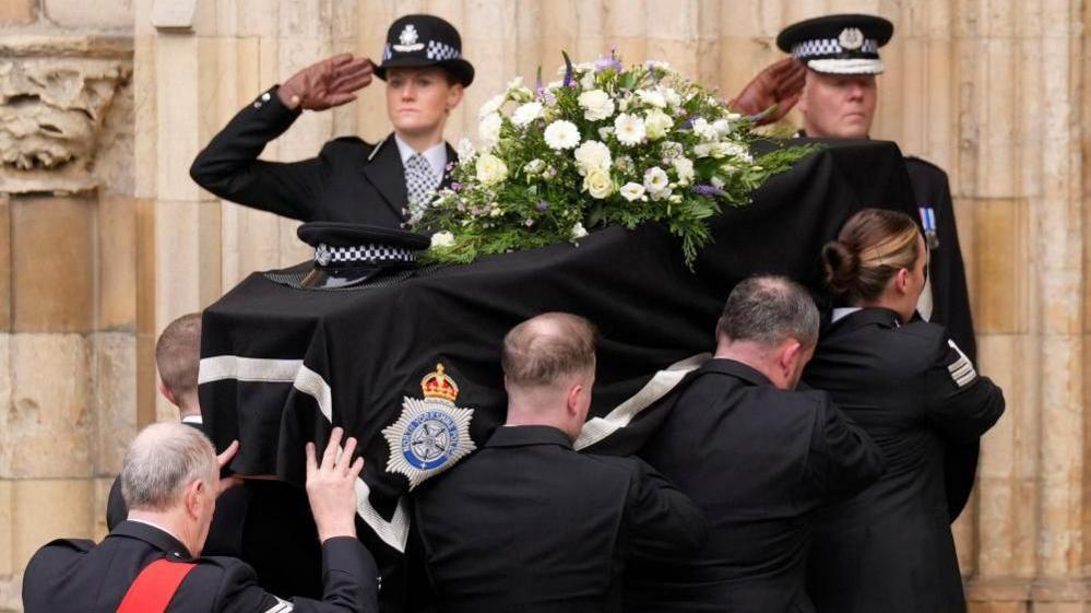 The coffin of police constable Rosie Prior is carried into York Minster for her funeral. Two police officers in uniform salute as the coffin is carried in, with black cloth over it and some flowers 