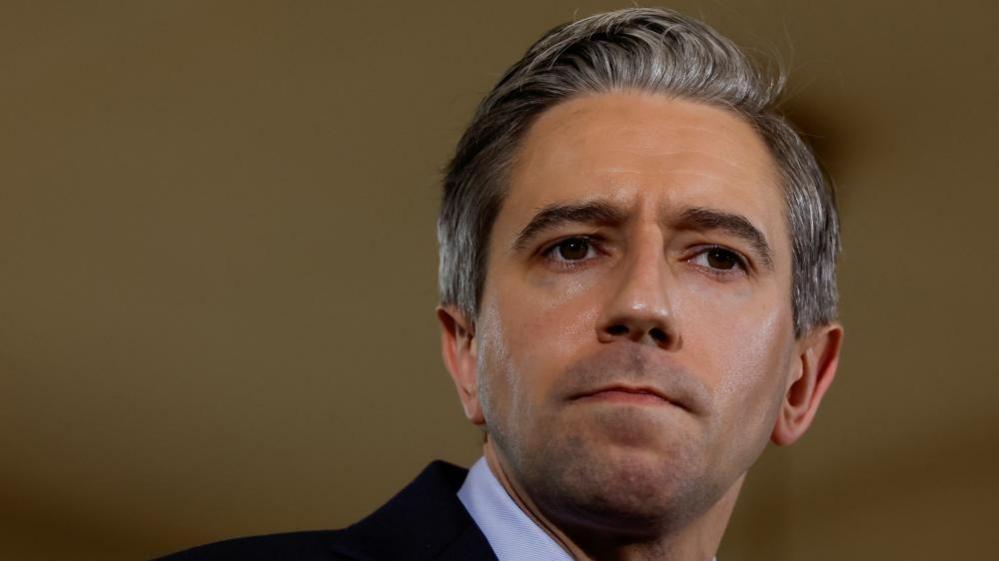 A man with short dark and grey hair with facial stubble, wearing a suit jacket and white shirt.