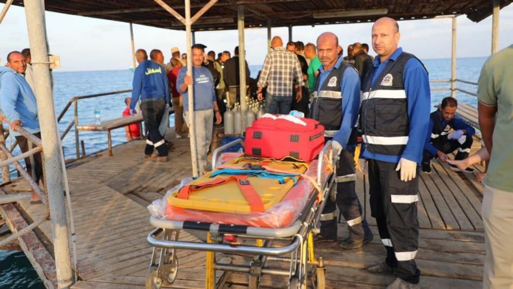 Medics stand beside a stretcher to wait for possible survivors after a boat sank at a harbour in Marsa Alam, Red Sea Governorate, in Egypt 25 Novermber 2024. Two medics are wearing black and blue medical suits. They are standing on a wooden platform by the sea.