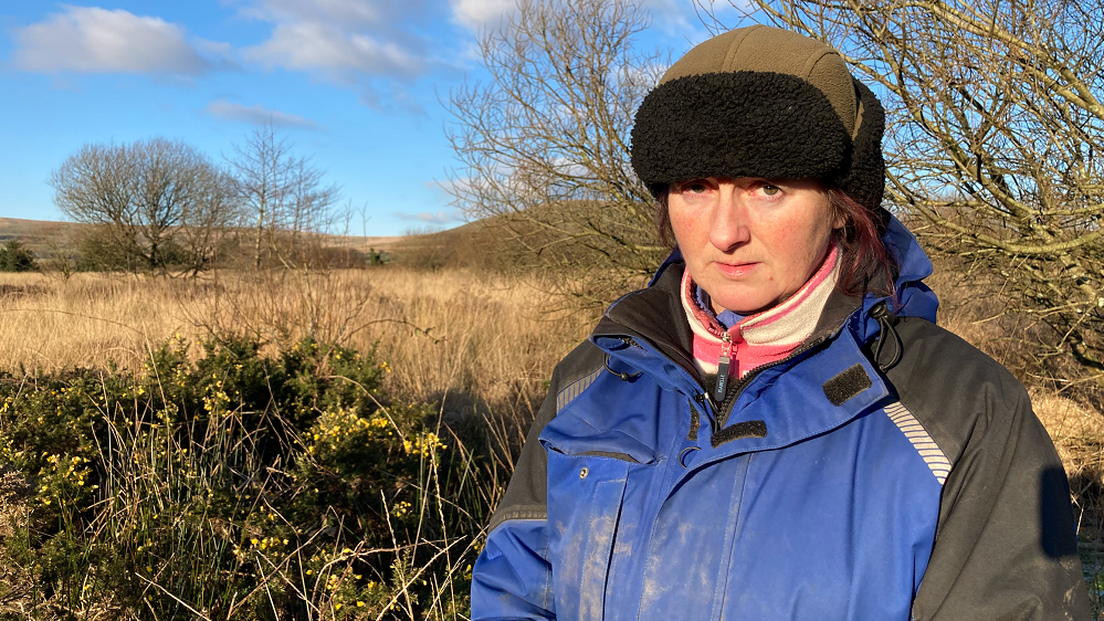 Photo of Anita in a hat and coat with trees and brambles around her