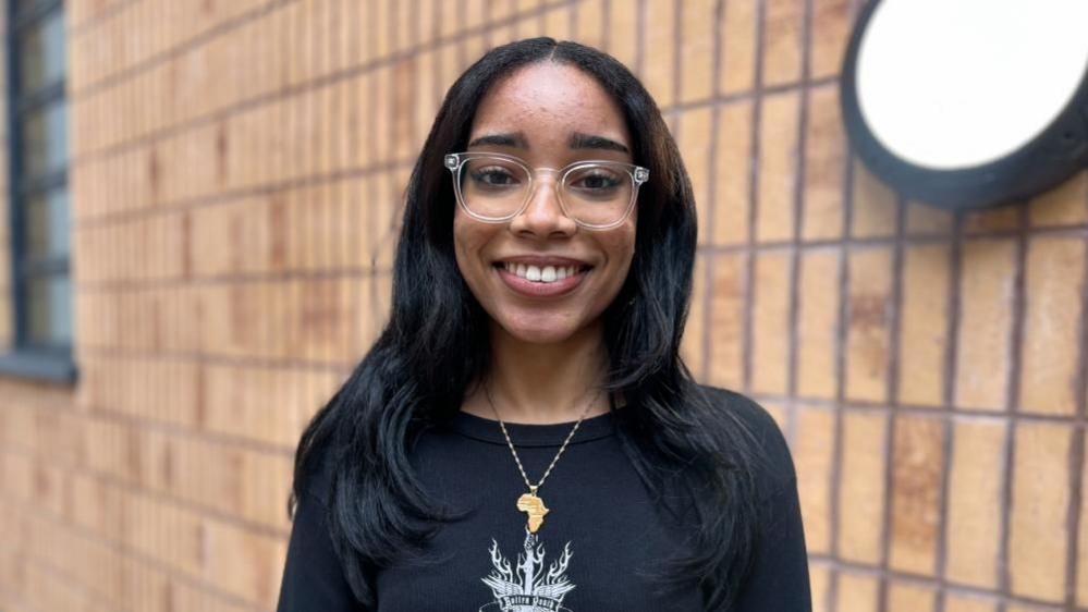 Sarah Dawelbeit, looking at the camera. She has transparent plastic glasses and black hair, and is wearing a necklace in the shape of the African continent.