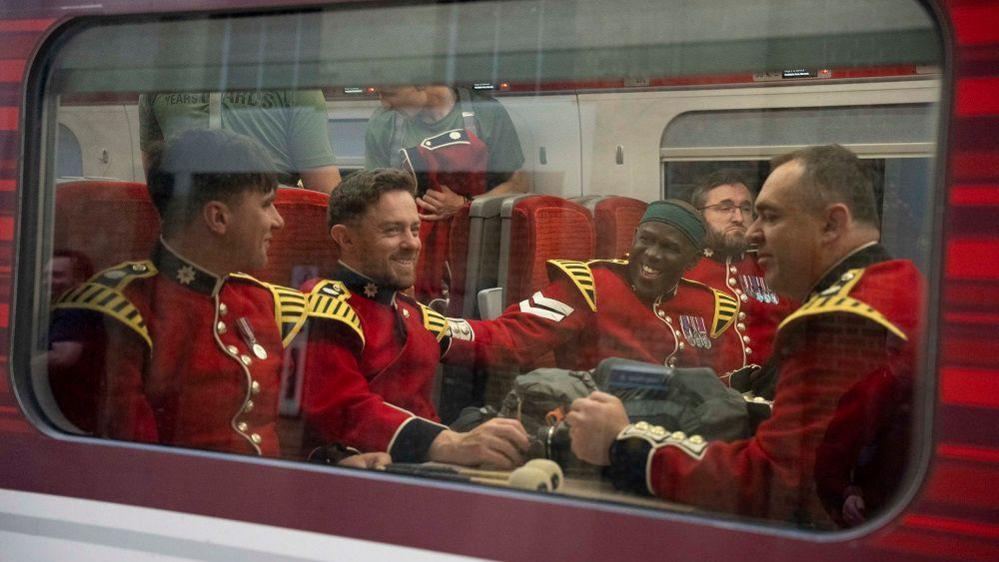 A group of guards sitting on a train with red seats. They are wearing the red uniform with yellow shoulders. They are smiling and chatting, one man has his had on the shoulder of another.