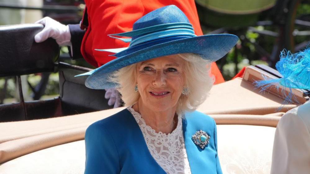 Queen Camilla arrives in a horse-drawn carriage wearing a blue suit and wide-brimmed hat