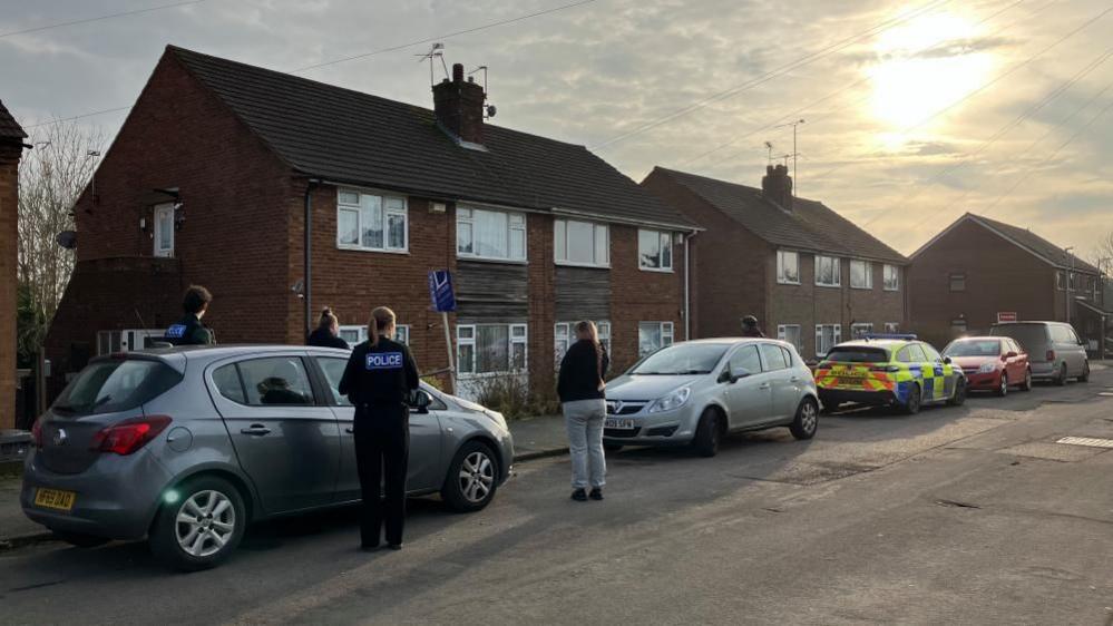 Police officers and members of the public stand on a residential street with cars, which includes a police vehicle, parked along the pavement.