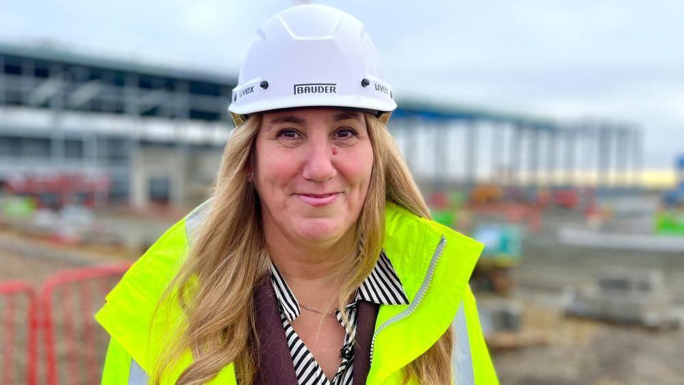 A blonde woman is wearing a hard hat and high visibility jacket. She is standing on a building site and you can see she is wearing a suit. The woman has long blonde hair. 